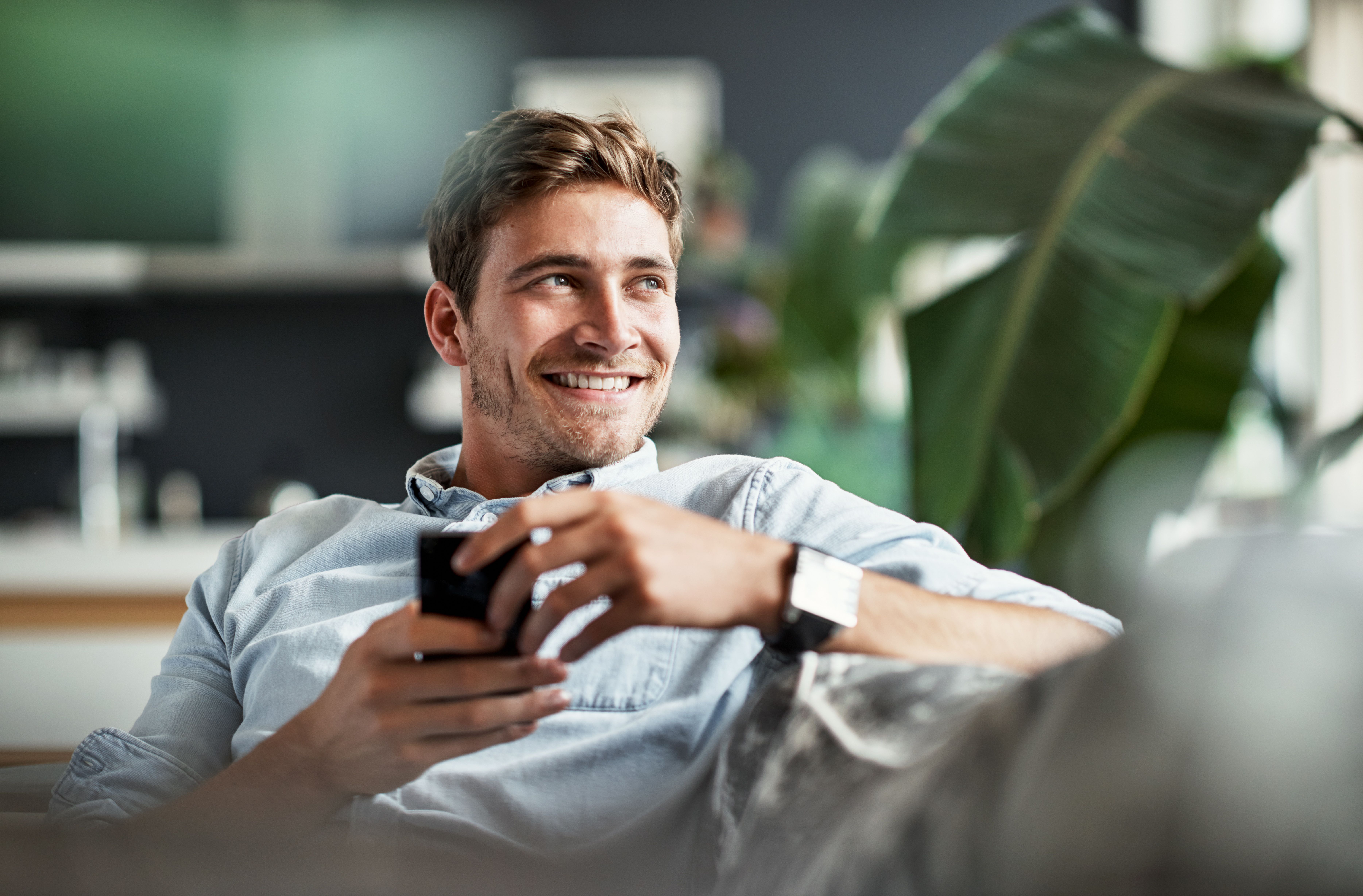 Young man in a denim shirt smiling and holding a smartphone while relaxing indoors