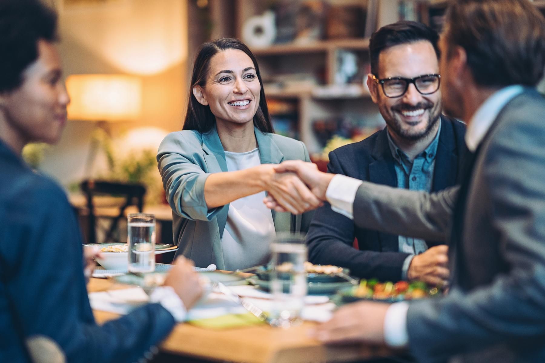 Two colleagues shaking hands and smiling across a table during a business meeting
