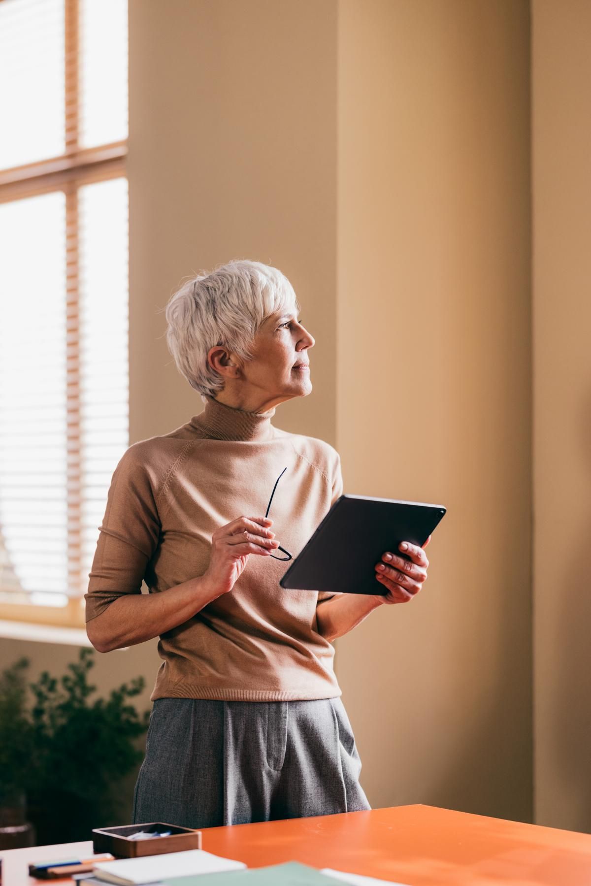 Professional older woman holding a tablet and glasses, looking out of an office window