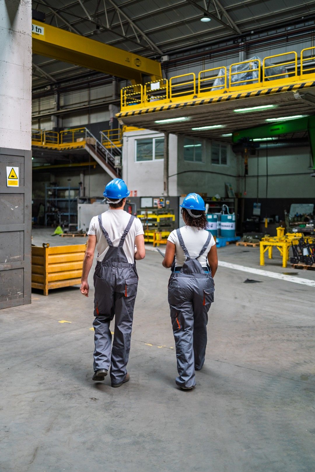 Two workers in blue hard hats and overalls walking through an industrial warehouse