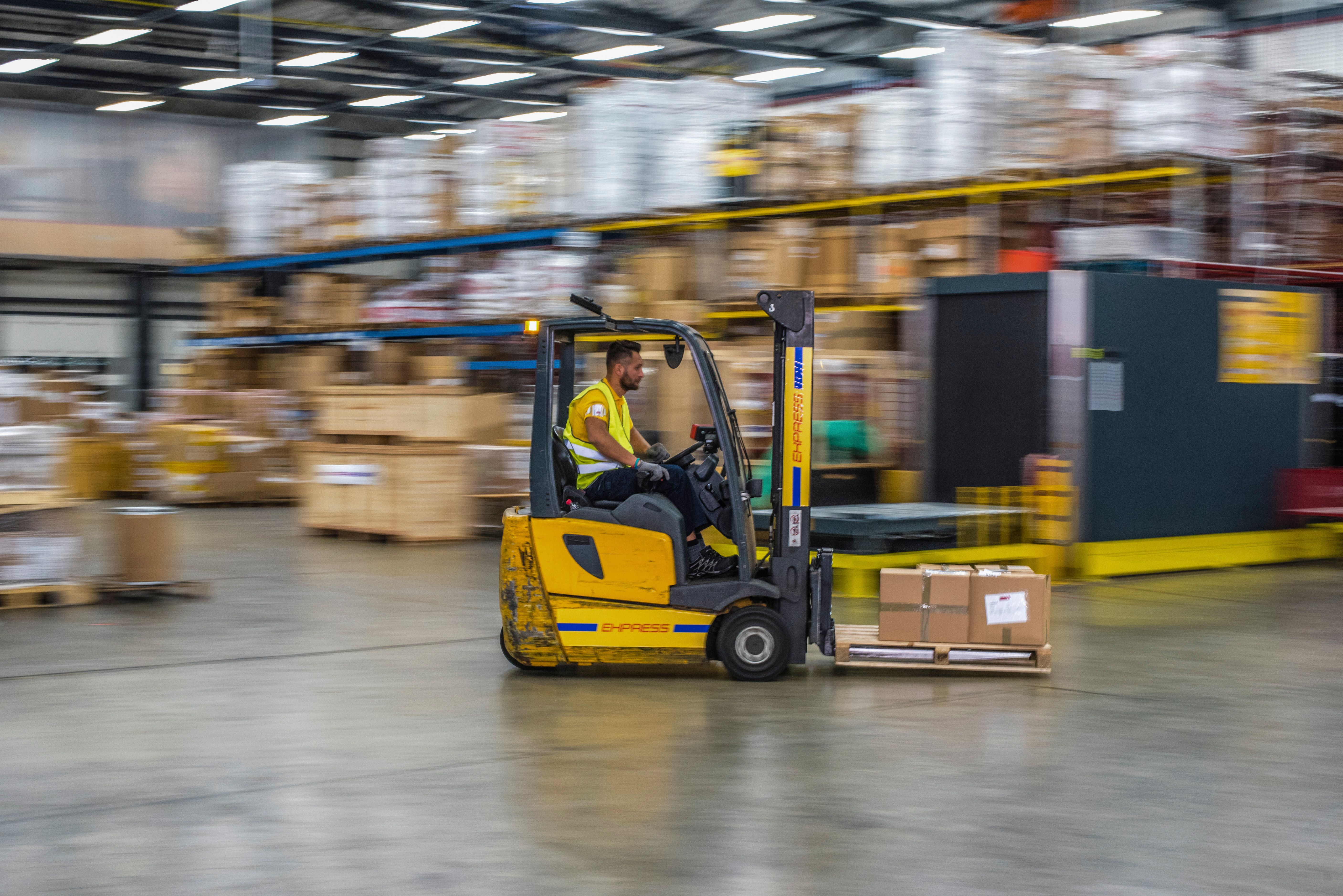 Operator driving a yellow counterbalance forklift through a busy warehouse