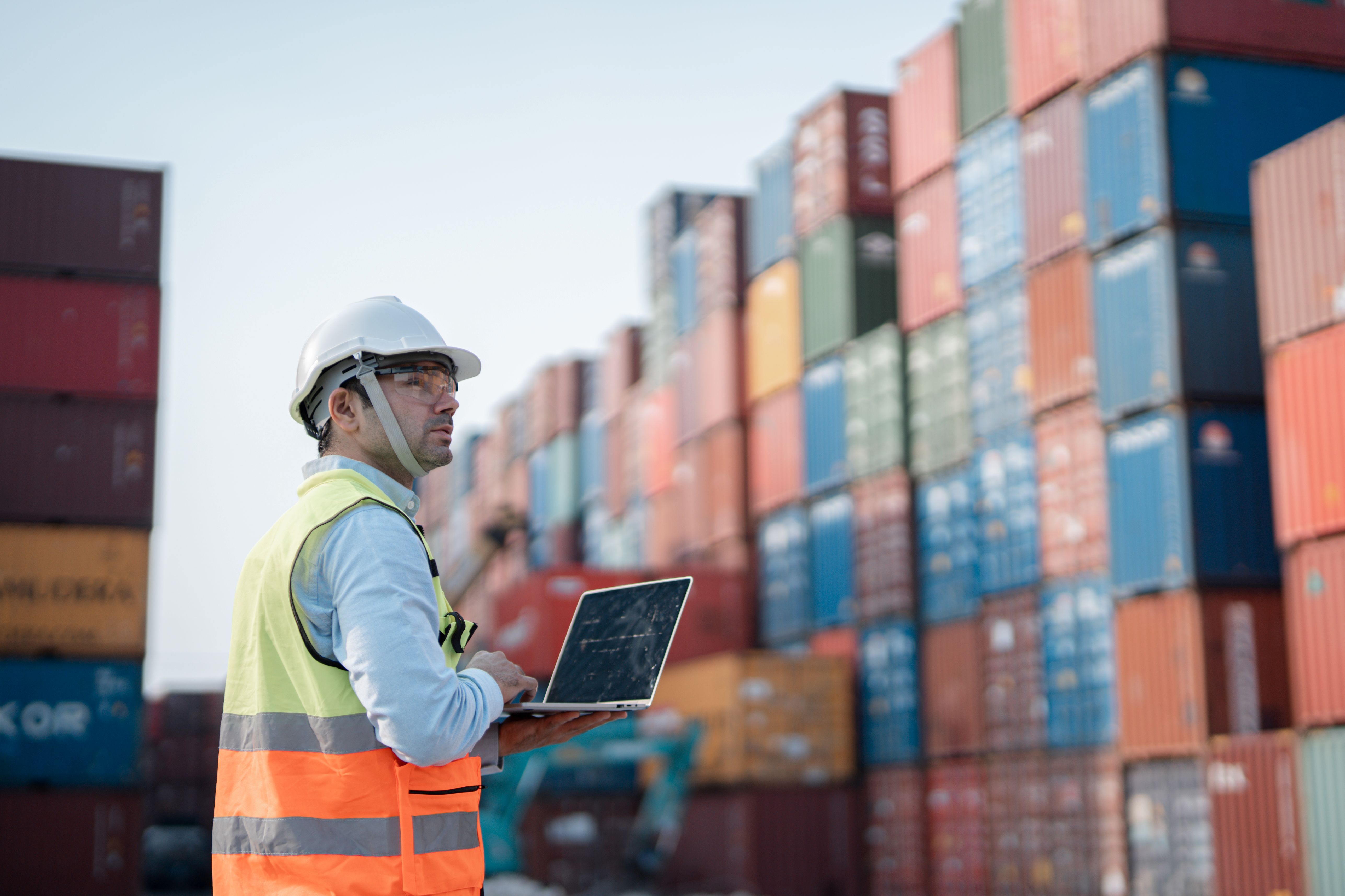 Worker in a hard hat and hi-vis vest reviewing data on a laptop in front of stacked shipping containers