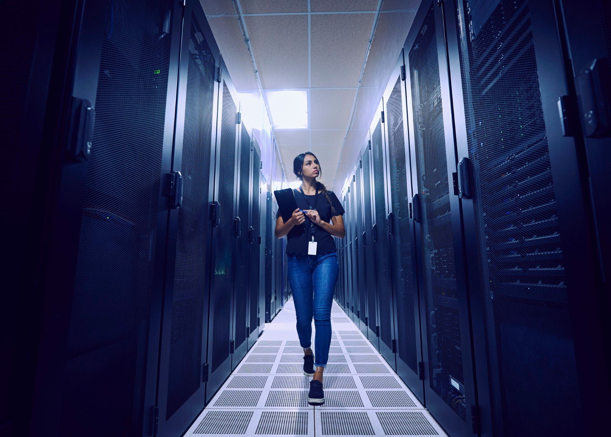 Female data center engineer with a lanyard walking down a narrow aisle between server racks