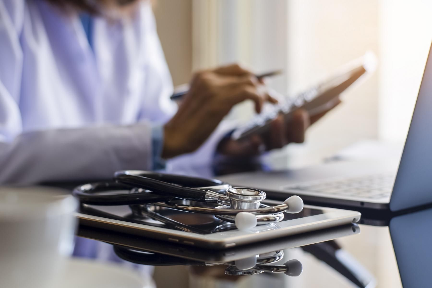 A clinician in a white coat using a calculator at a desk beside a stethoscope and tablet.
