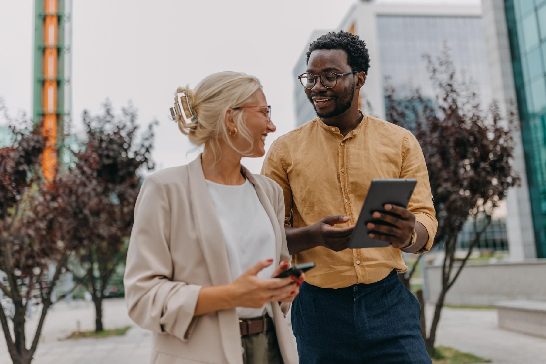 Two colleagues walking outdoors in a city, talking while one holds a tablet and the other a phone.
