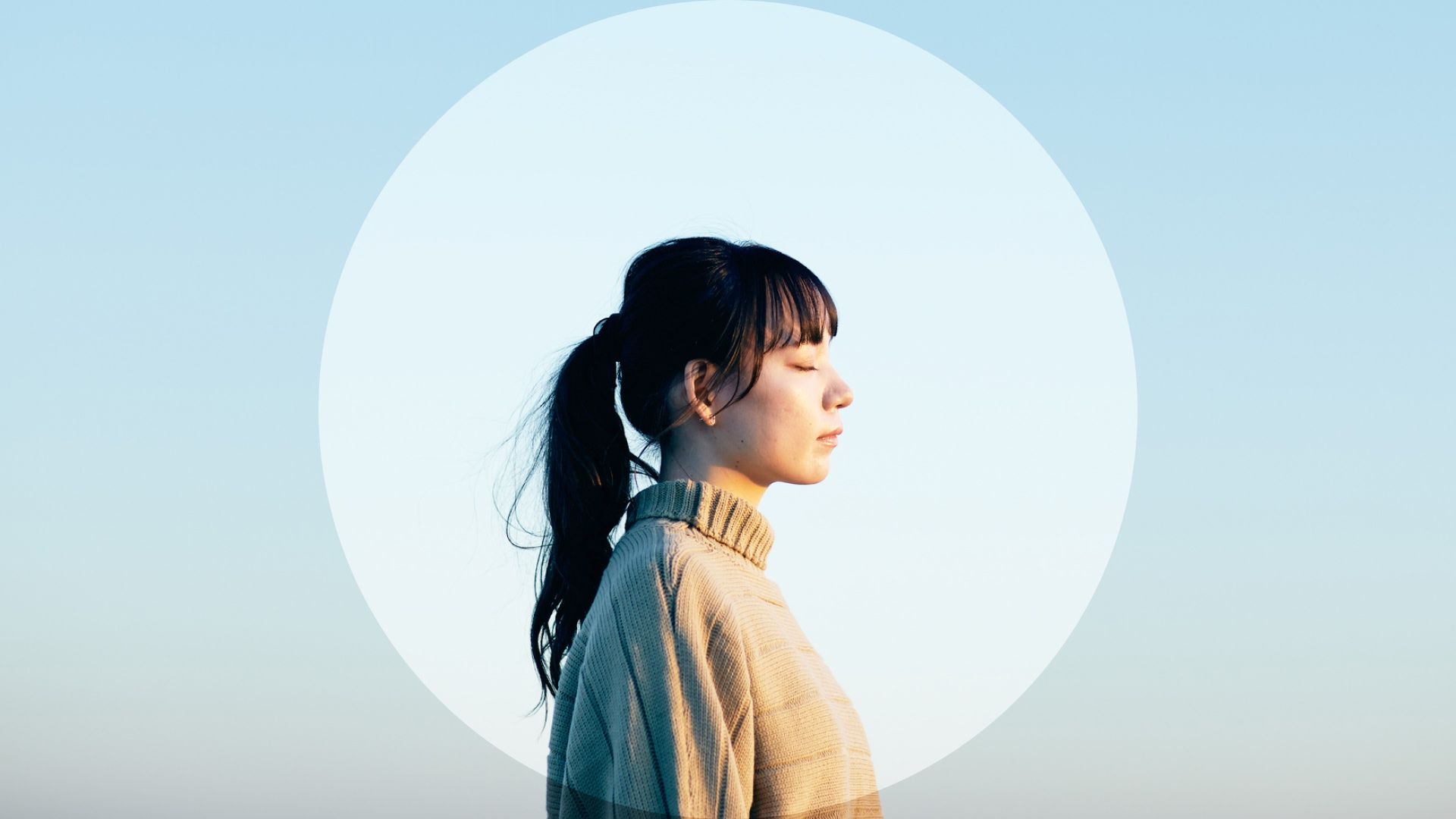 Woman in profile with her eyes closed, lit by sunlight against a pale blue sky.