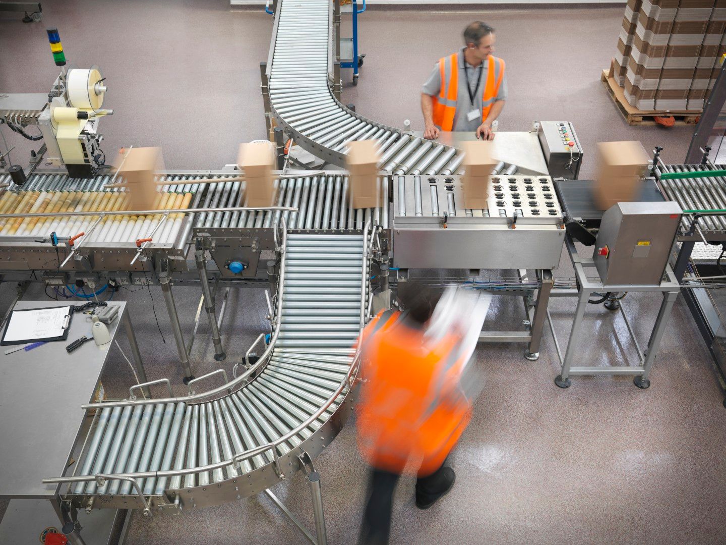 Overhead view of a warehouse conveyor belt system moving cardboard boxes, with two workers in orange safety vests operating the equipment.