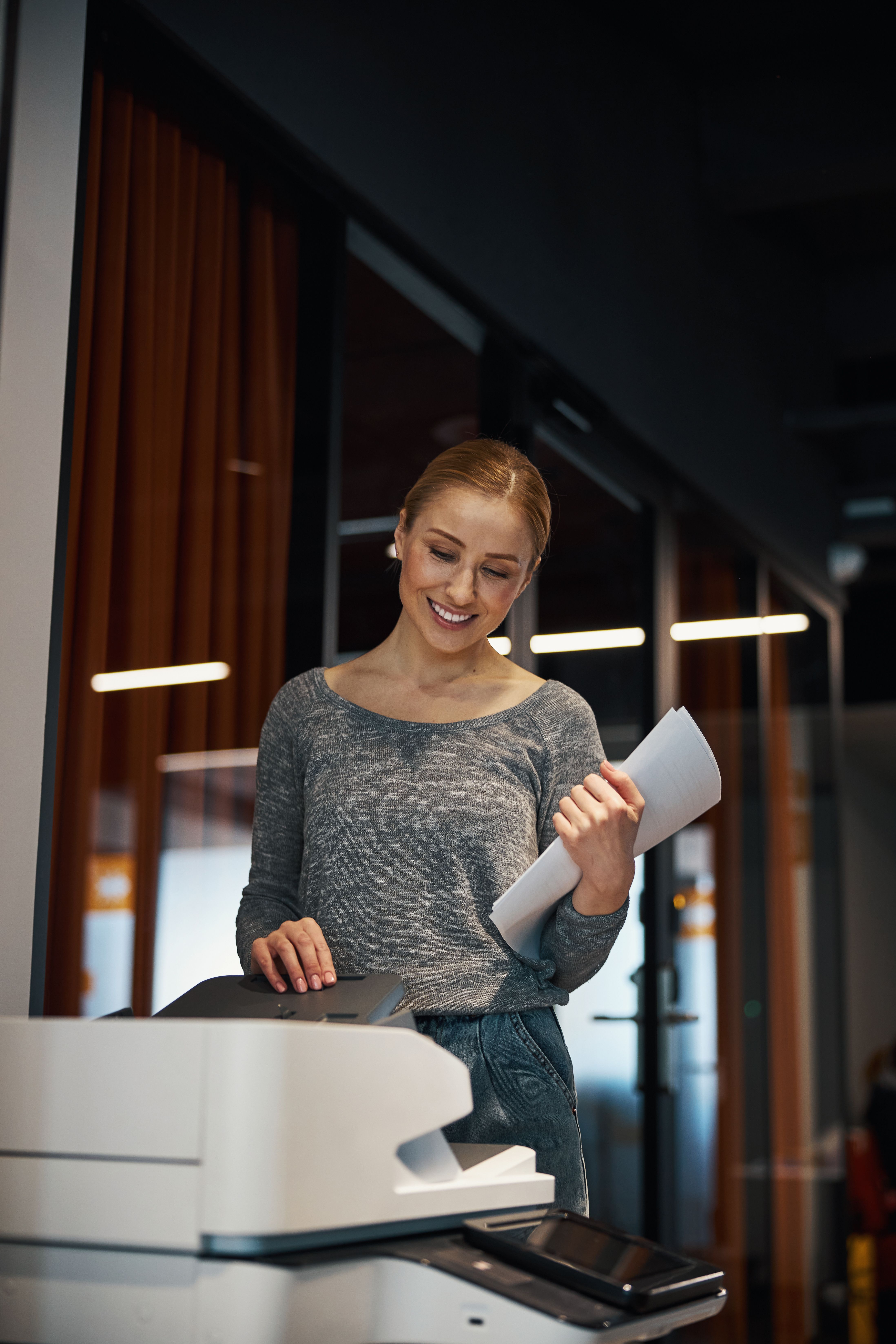 Smiling woman with blonde hair holding printed pages beside an office printer