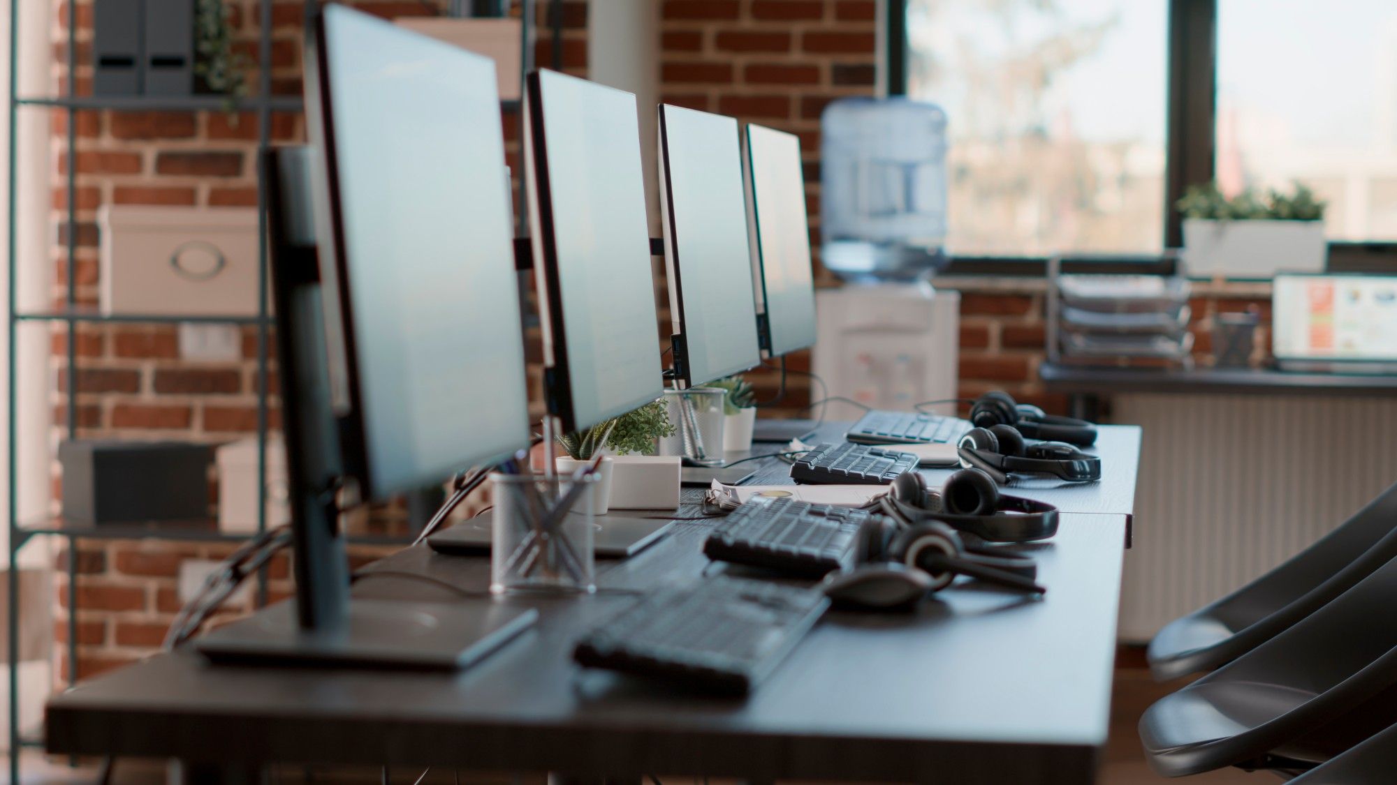 Row of desktop computer monitors, keyboards, and headsets on a long shared desk beside an exposed brick wall